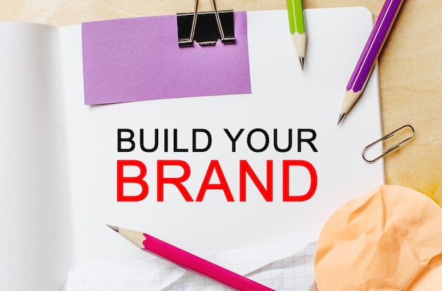 Text Build Your Brand on a white note background with pencils, stickers and paper clips