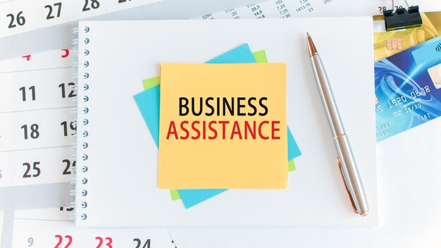 Text business assistance written on yellow paper square shape. Credit cards, pen, stationery on the white desktop background. Business, finance and education concept. Selective focus.
