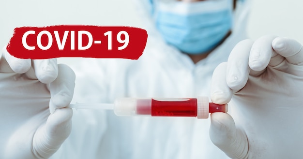 빨간색 경고 표시에 covid-19라는 텍스트를 표시합니다. 의사의 손에 코로나바이러스 혈액 검사. 보호용 의료복을 입은 의사, 생물학적 위험, 흰색 바탕에 마스크. 의학 실험실 건강 관리의 개념입니다.