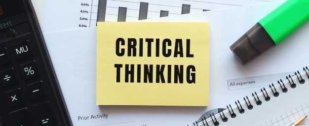 Text CRITICAL THINKING on the page of a notepad lying on financial charts on the office desk