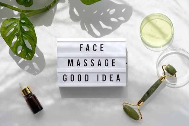 Text Face Massage Good Idea on light box. Lightbox on Moisturizer, green jade face roller with exotic leaves. Monstera Adansonii leaves. Sunshine, long shadows. Top view on off white background.