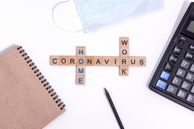 Text from wooden letters coronavirus work from home. Office supplies, a calculator, paper Notepad for notes, a pencil and a protective medical mask on desktop.