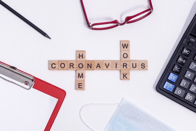 Text from wooden letters coronavirus work from home. Office supplies, calculator, tablet, glasses, pencil and protective medical mask on the desktop.