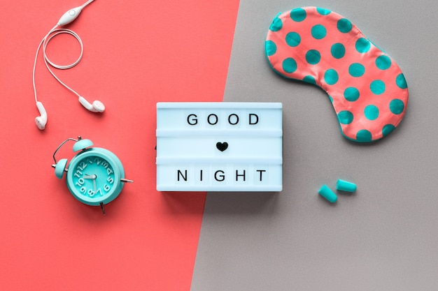 テキスト「おやすみ」健康的な夜の睡眠の創造的なコンセプト。水玉模様のスリーピングマスク、目覚まし時計、イヤホン、耳栓。フラット横たわっていた、トップビュー、2つのトーンのサンゴとターコイズブルーの紙の壁を分割します。