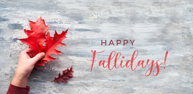 Text Happy Fallidays. Flat lay, top view, hand holding twig with red oak leaves on textured grey wood