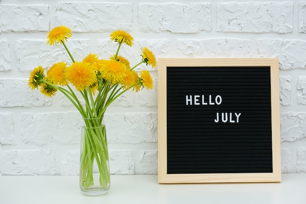 Text Hello July on black letter board and bouquet of yellow dandelions flowers in vase on background white brick wall.