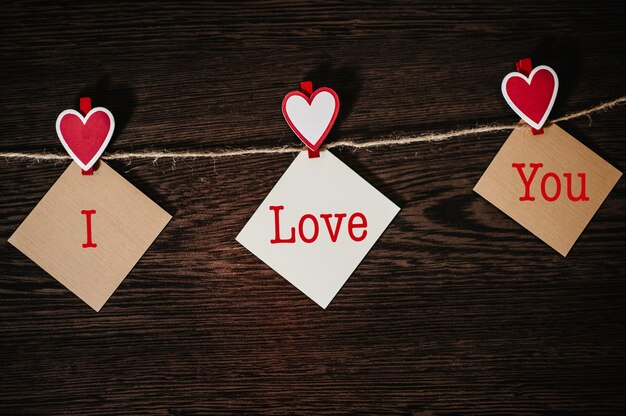 Text I love you. Craft paper, three blank, with red heart hanging on rope on brown wooden wall