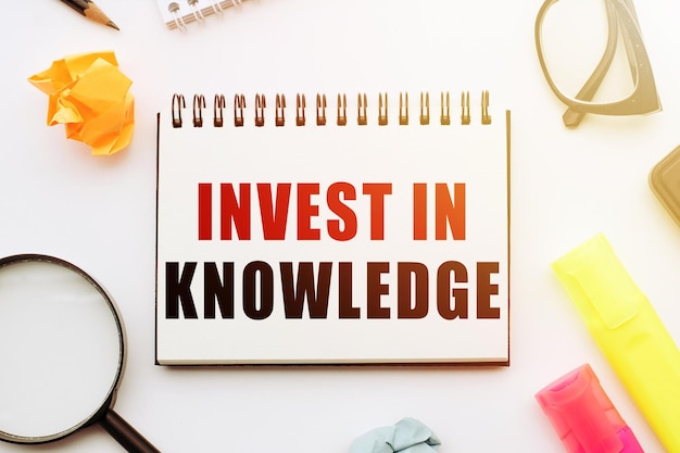 Text INVEST IN KNOWLEDGE in notebook on white table with office tools