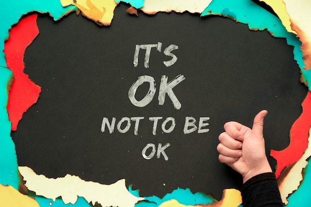 Text "It's OK not to be OK " in burned paper frame on black wall and hand showing OK sign. Positive quote for persons suffering from personality disorder and Mental Health Awareness Month.