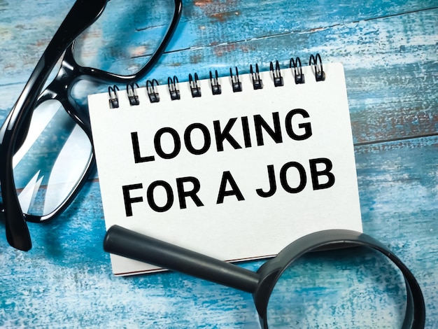 Text LOOKING FOR A JOB written on notebook with magnifying glass and glasses on blue wooden background