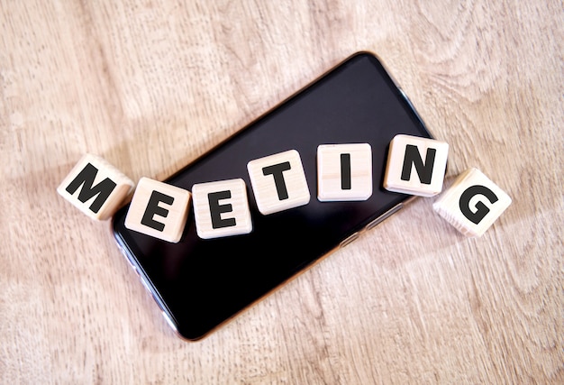 Text MEETING on wooden cubes on a black smartphone. Cubes lay on a black smartphone on the wooden table.