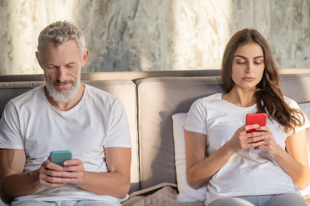 Текстовое сообщение. Обеспокоенный глядя бородатый взрослый мужчина и молодая длинноволосая женщина со своими смартфонами, сидя на кровати