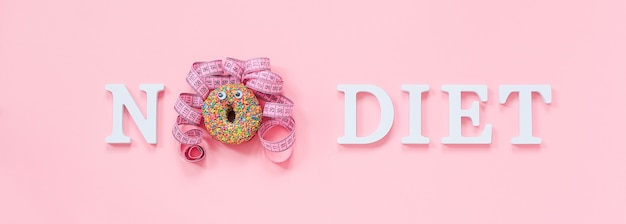 Photo text no diet and abstract funny face of woman from donut with eyes and hair from centimeter tape on plate on pink wall