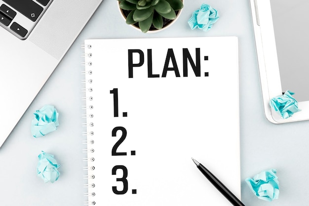 Text PLAN on note. Laptop, pieces of paper, pen and plant on office desk. Flat lay, top view. Planning concept.