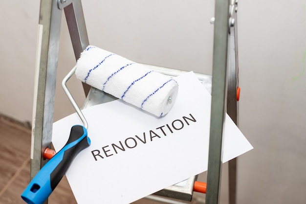 text renovation, black letters on white paper. home improvement, dirty paint ladder, roller, brush and tray.repairing room.Stepladder and different tools in room. Interior renovation