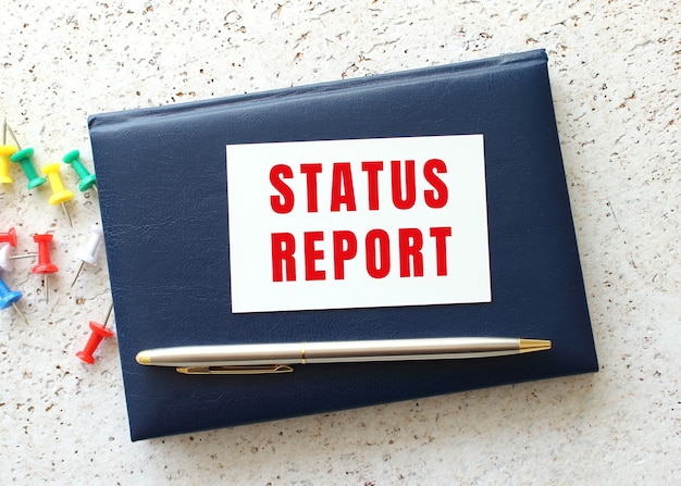 Text STATUS REPORT on a business card lying on a blue notebook next to the pen