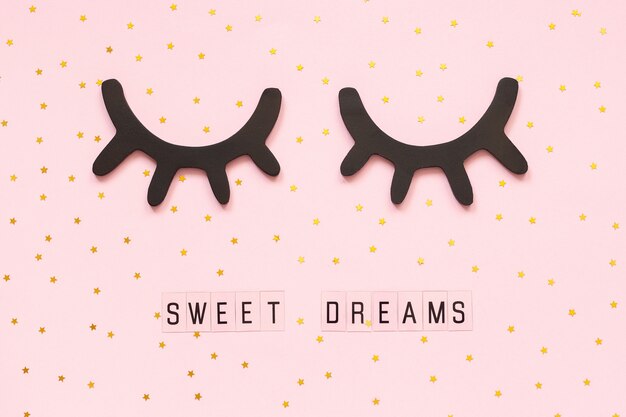 Text Sweet dreams and decorative wooden black eyelashes, closed eyes gold star on pink background. 