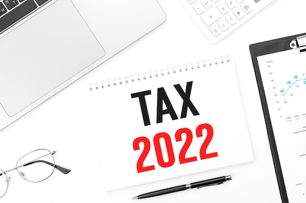 Text TAX 2022 on card. Laptop, glasses, pen, calculating machine and clipboard with charts and graphs. Business plan. Top view.