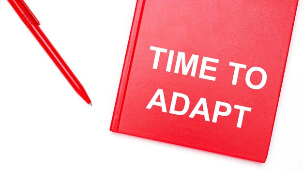 The text TIME TO ADAPT is written on a red notepad near a red pen on a white table in the office Business concept