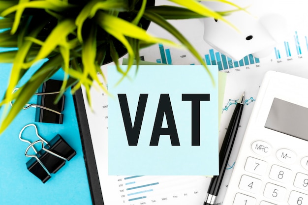 Text vat - value-added tax on blue sticker, calculator, pen, piggy, graphs, plant. business flat lay