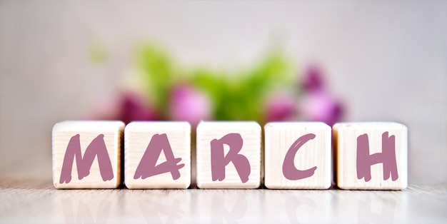 Text of the word MARCH on wooden cubes by light wooden table. Flowers in the background