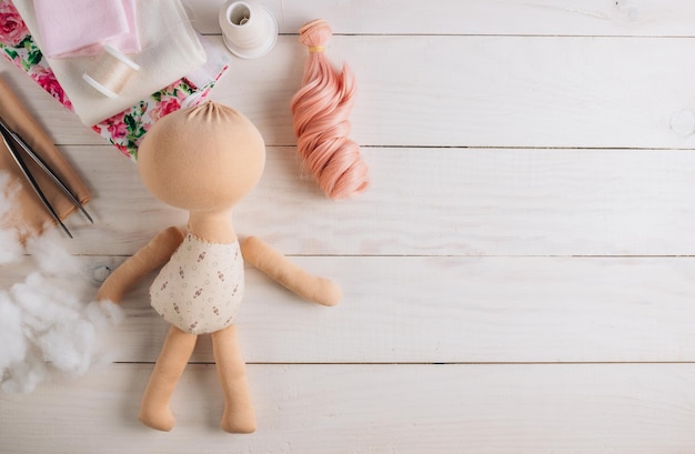 Textile doll in the sewing process with accessories and materials fabric thread needle on white wooden background Top view with copy space Flat lay