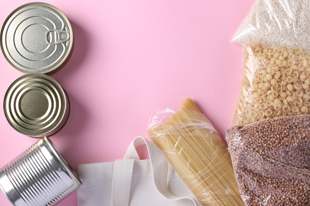 Textile Grocery Bag with food supplies crisis food stock on pink surface. Rice, buckwheat, pasta, canned food. Donation food