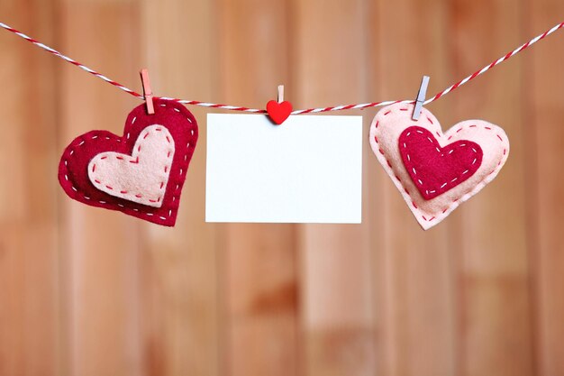 Textile hearts and empty sheet hanging on cord against wooden background