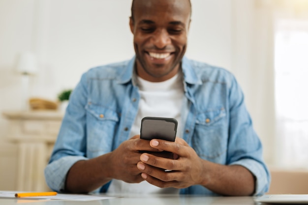 Текстовые сообщения. Привлекательный счастливый темноволосый афро-американский мужчина улыбается и пишет сообщение на своем телефоне, сидя за столом