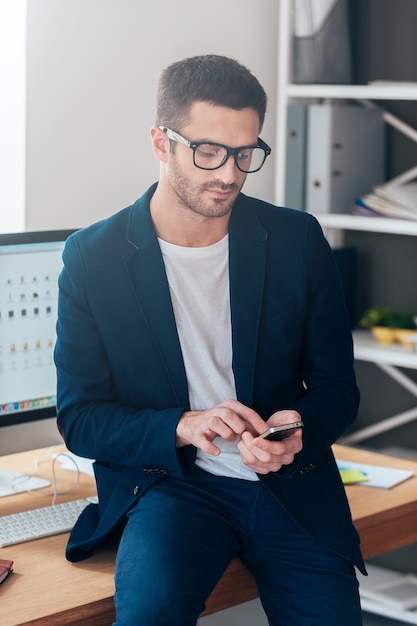 Photo texting business message. confident young man holding smart phone and looking at it while leaning at the office desk