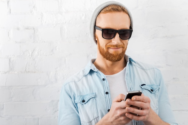 Texting to friend. Handsome young bearded man holding mobile phone and smiling while standing against brick wall