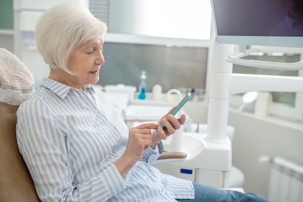 テキストメッセージ。歯科医院に座ってスマートフォンを使用している年配の女性