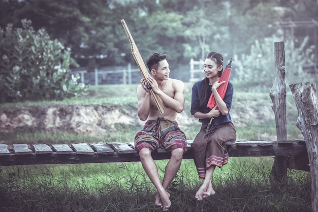 Thai couple on traditional clothes with umbrellas sitting on a wooden bridge