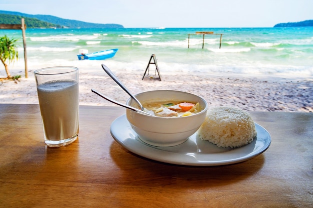 タイ料理。美しい海の風景を背景に、テーブルの上にココナッツミルクとご飯が入ったスープのプレート。ビーチレストランでの朝食。カンボジア、コーロンサムレム島の海岸。