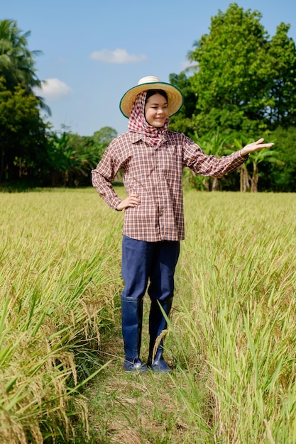 전통 셔츠와 모자를 쓴 태국 여성 농부가 들판 한가운데 제품을 들고 서서 포즈를 취하고 있습니다.