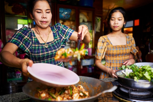 Thai mother and daughter plating freshly cooked red curry in rustic traditional kitchen