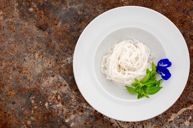 Vermicelli tailandesi spaghetti di riso tailandesi kanom jeen con foglie di basilico peloso e fiori di pisello farfalla in piatto di ceramica bianca su sfondo texture arrugginita copia spazio per testo vista dall'alto piatto cibo tailandese