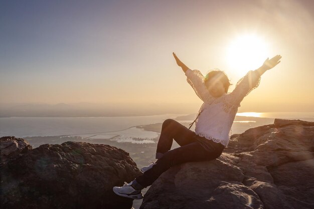 Photo thai woman sitting on a rock on the mountain raise your hands happy amidst beautiful views and the sunrise background in the morning freedom and travel concept