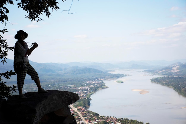 Thai woman travel visit and posing take photo on ridge stone of cliffs at Wat Pha Tak Suea temple with view of landscape of Nongkhai city and loas and Mekong river in Nong Khai Thailand