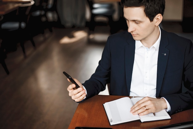 Thank you,for making me part Handsome young male brunette holding a smartphone looking at the screen while sitting on a desk with a hand on a notebook.