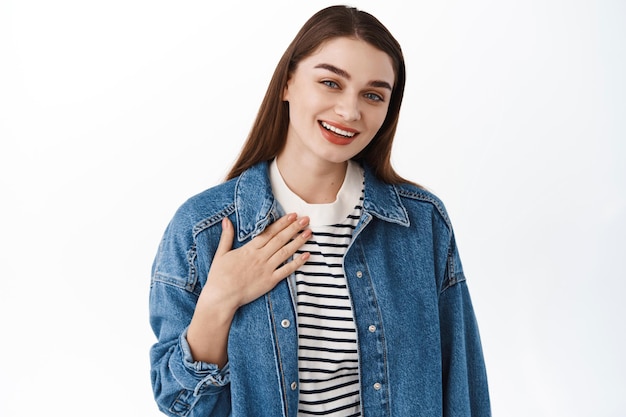 Thank you. Smiling attractive woman looks happy and flattered, holding hands on her chest, introduce herself, express gratitude, standing over white background