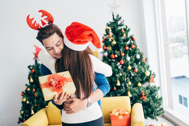 驚きのクリスマス プレゼントを持つ感謝のアジア人自宅で妻を抱き締める新年を祝うクリスマス イブの伝統として贈り物を提示彼女のガール フレンドからお祝いの言葉を受け取る幸せなアジア人男性