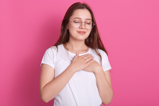 thankful young woman wearing white casual t shirt posing isolated over pink, standing with closed eyes, keeps hands at heart chest, feels gratitude, grateful.