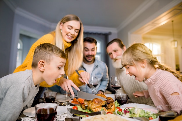 Thanksgiving Celebration Tradition Family Dinner Concept. Funny photo of big family sitting at the table, mother is cutting turkey and two little excited children looking at the food