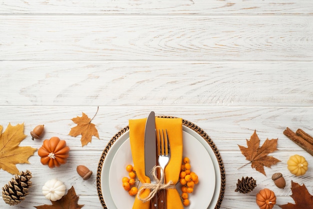 Photo thanksgiving day concept top view photo of plate cutlery fork knife napkin rowan maple leaves pine cones acorns pumpkins and cinnamon sticks on isolated white wooden table background with copyspace