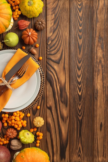 Thanksgiving day concept Top view vertical photo of plate knife fork napkin vegetables pumpkins pattypans pear walnut acorn physalis and rowan on isolated dark wooden table background with copyspace