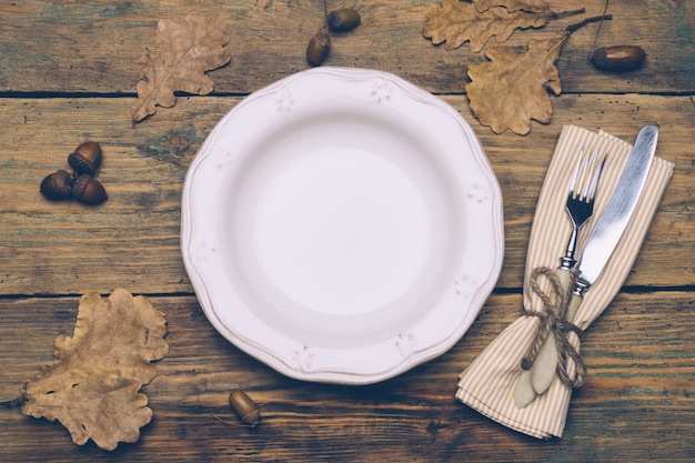 Фото Фон концепции обеда благодарения: белая тарелка, ложка и вилка на деревянном деревенском столе с осенними листьями, красными ягодами и желудями