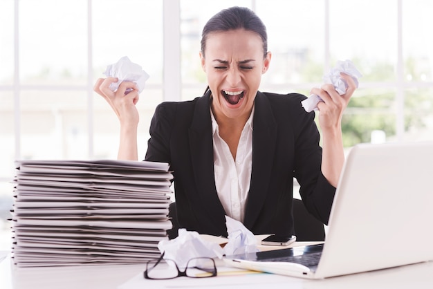 That is too much for me! Furious young woman in formalwear holding papers in hands and shouting while sitting at her working place