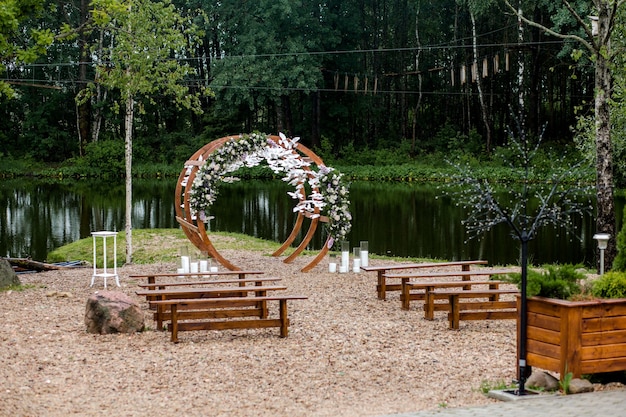 사진 부두의 강 근처 숲에서 결혼식 장소. 꽃, 녹지, 흰 새, 양초, 손님을위한 의자 벤치로 장식 된 나무 둥근 아치. 귀엽고 세련된 장식