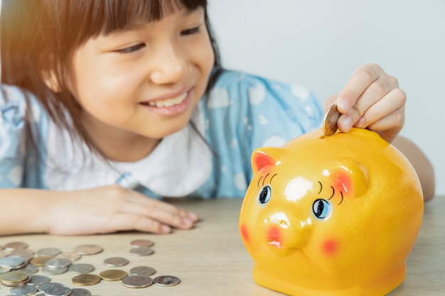 写真 アジアの女の子は白い壁でお金を節約するために貯金箱を喜んで落とします。お金の概念を保存します。